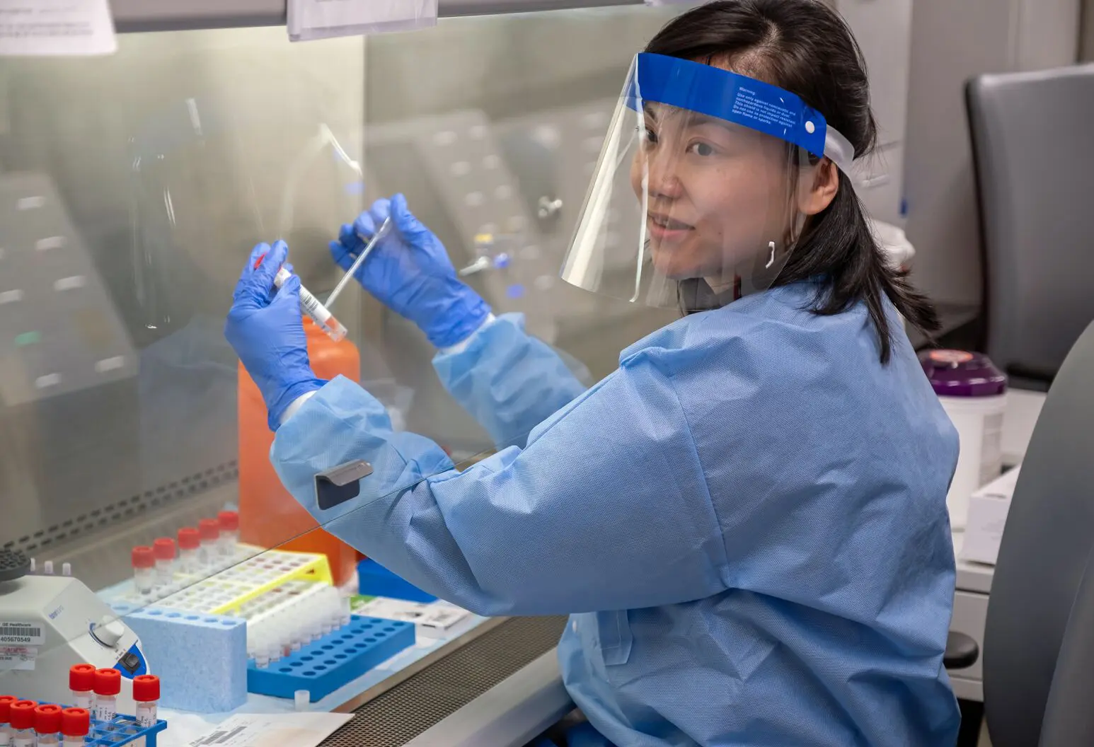 A woman in blue lab coat and gloves working with a test tube.